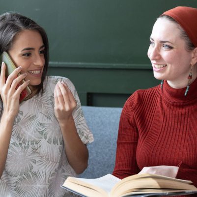 Two women sitting on couch talking on phone and reading