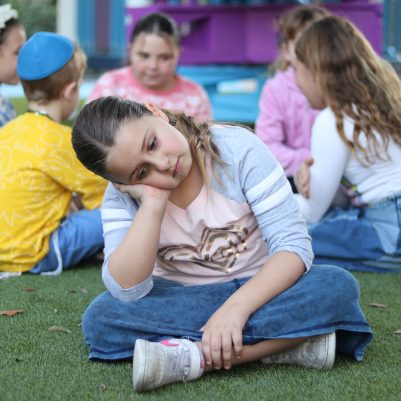 Sad Young Girl Sitting Alone on Grass While Other Children Play in Background
