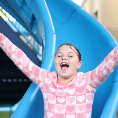 Happy Young Girl Sliding Down Playground Slide with Arms Raised