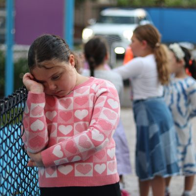 Crying Young Girl Leaning on a Playground Fence While Other Children Play